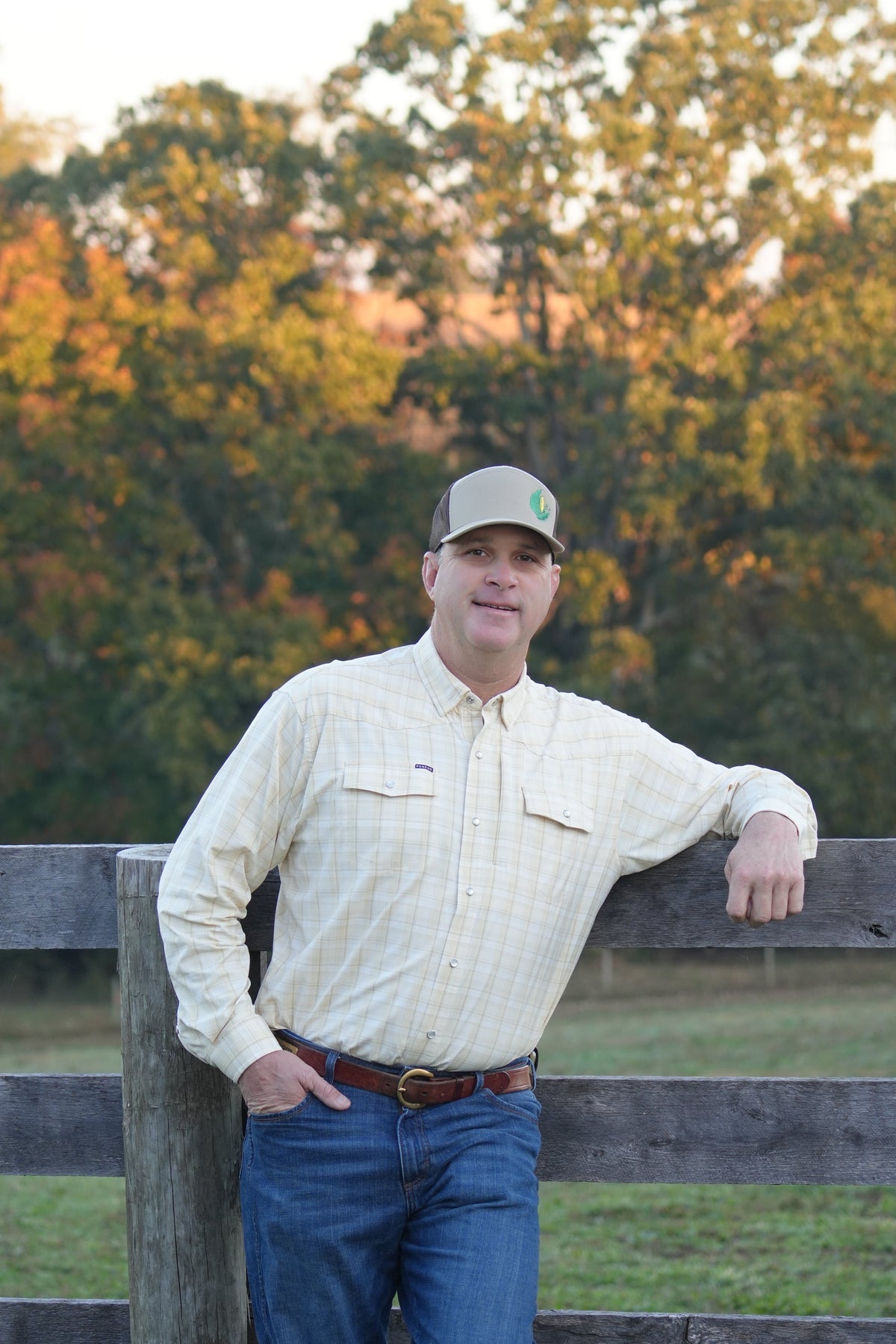 A man wearing a cream-colored long-sleeve button-up shirt, blue jeans, and a baseball cap leans against a wooden fence outdoors with trees in the background.