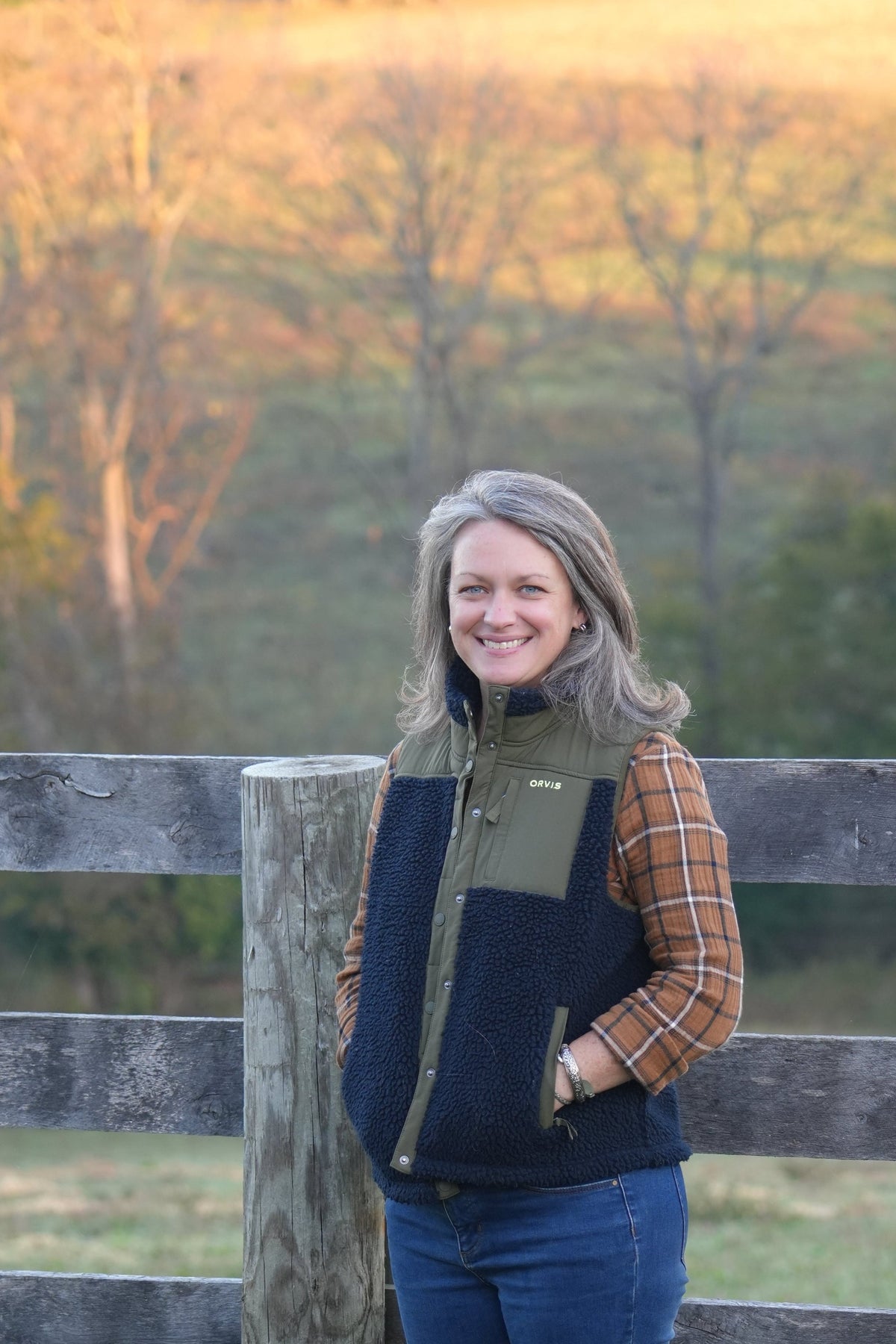 A woman stands outdoors beside a wooden fence, wearing a navy fleece vest over a brown plaid flannel shirt and blue jeans. The background shows a softly blurred rural landscape with trees and grass.